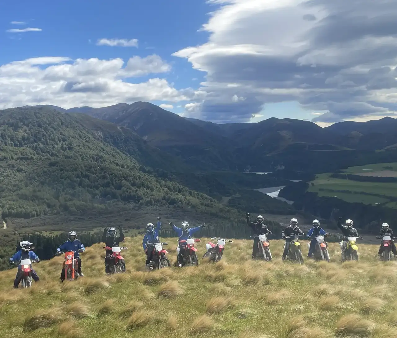 Twelve riders lined up along a tussock ridge above a river valley, several with an arm raised