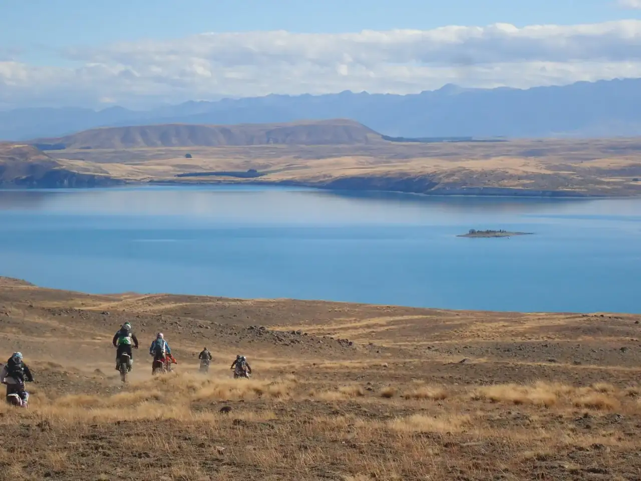 Riders crossing open tussock flats above a pale blue high country lake